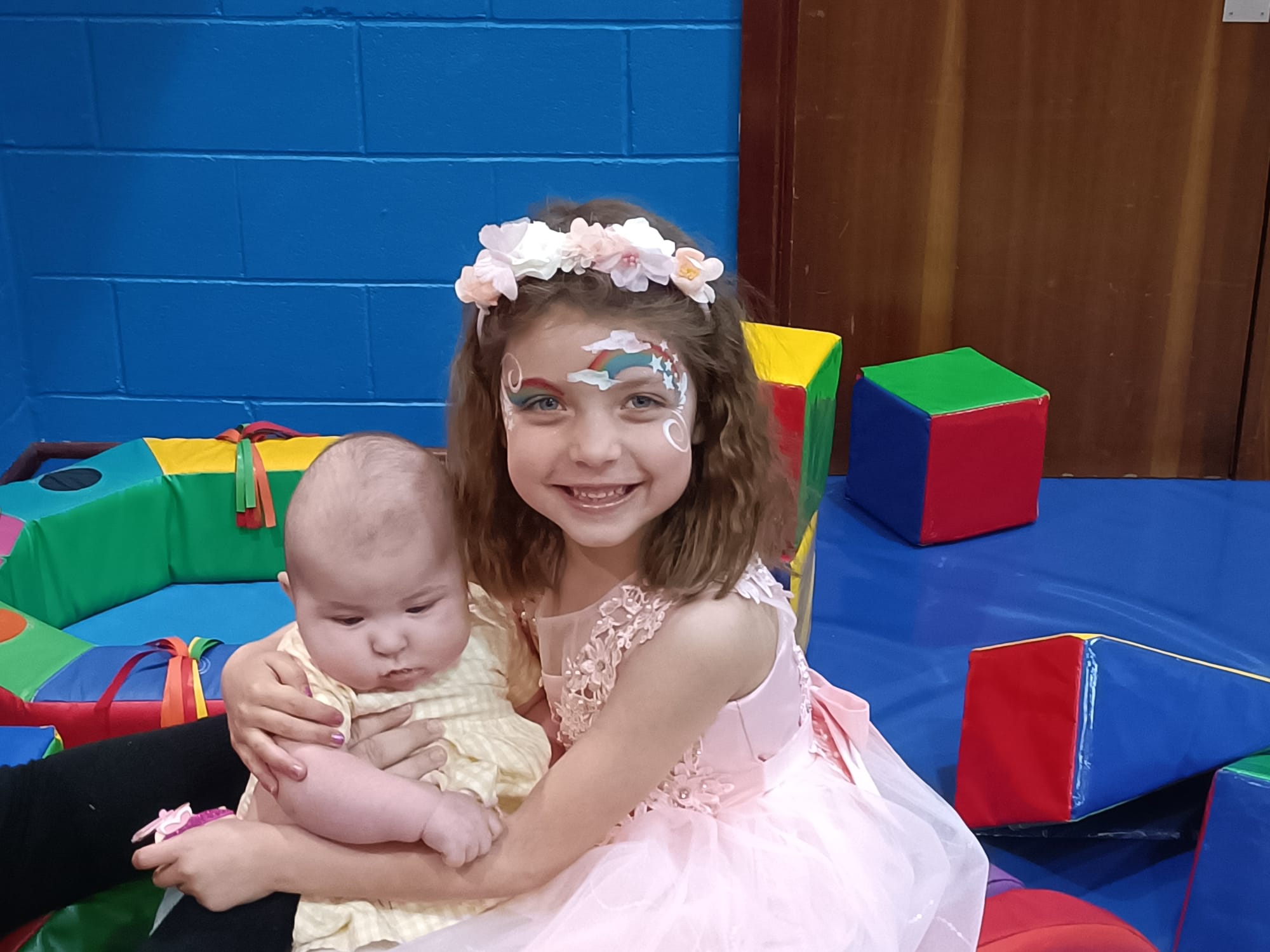 little girl with rainbow face paint holding a baby playing in soft play